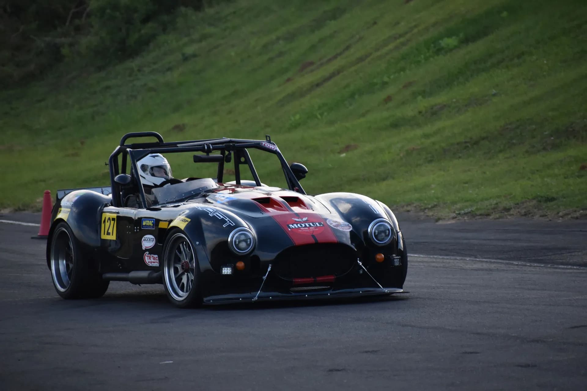 Black and red open-cockpit race car with driver driving on an asphalt track next to a green grassy slope.