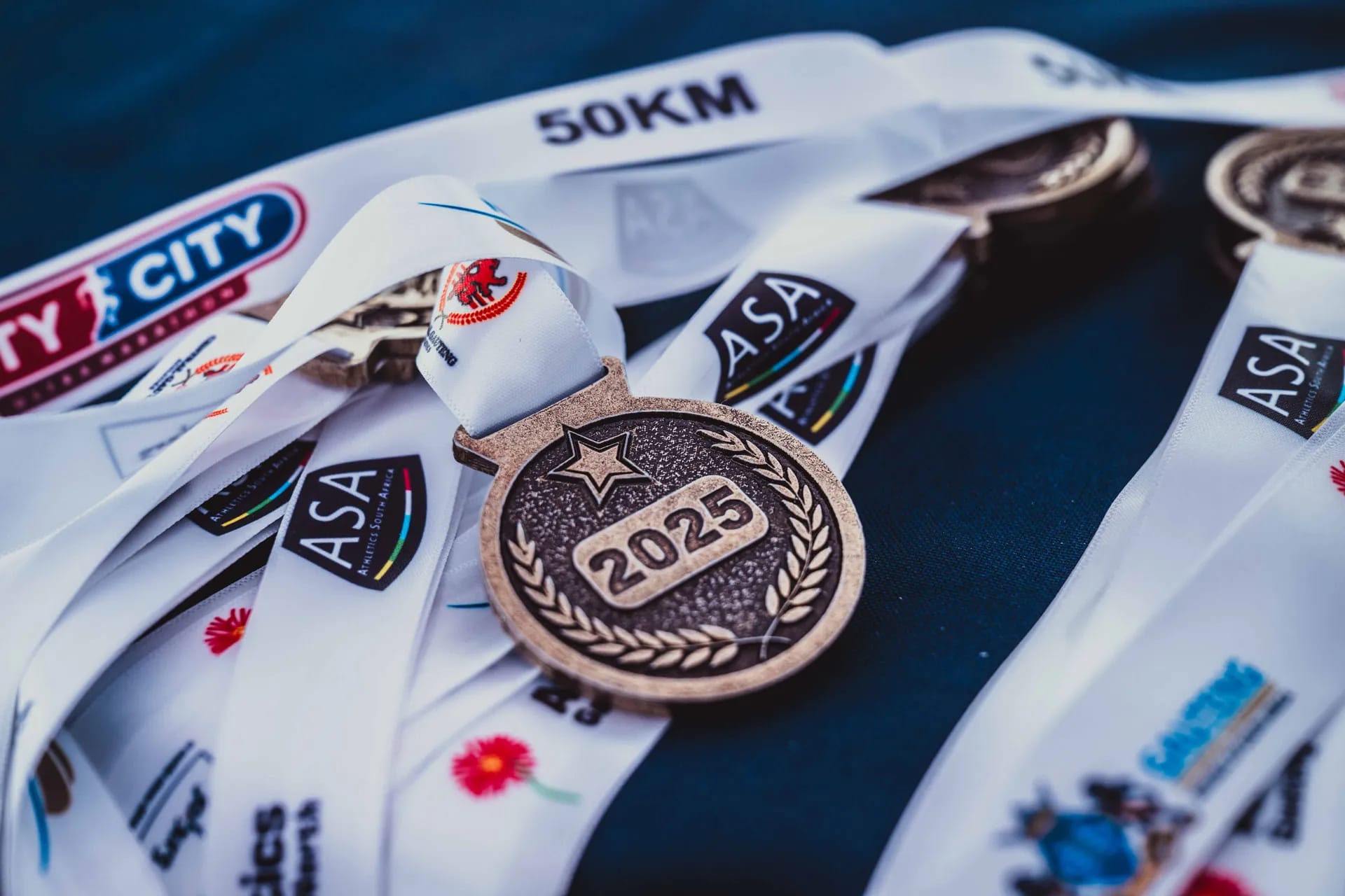 Bronze 2025 finisher medals with white ASA and 50km ribbons for the City to City marathon.