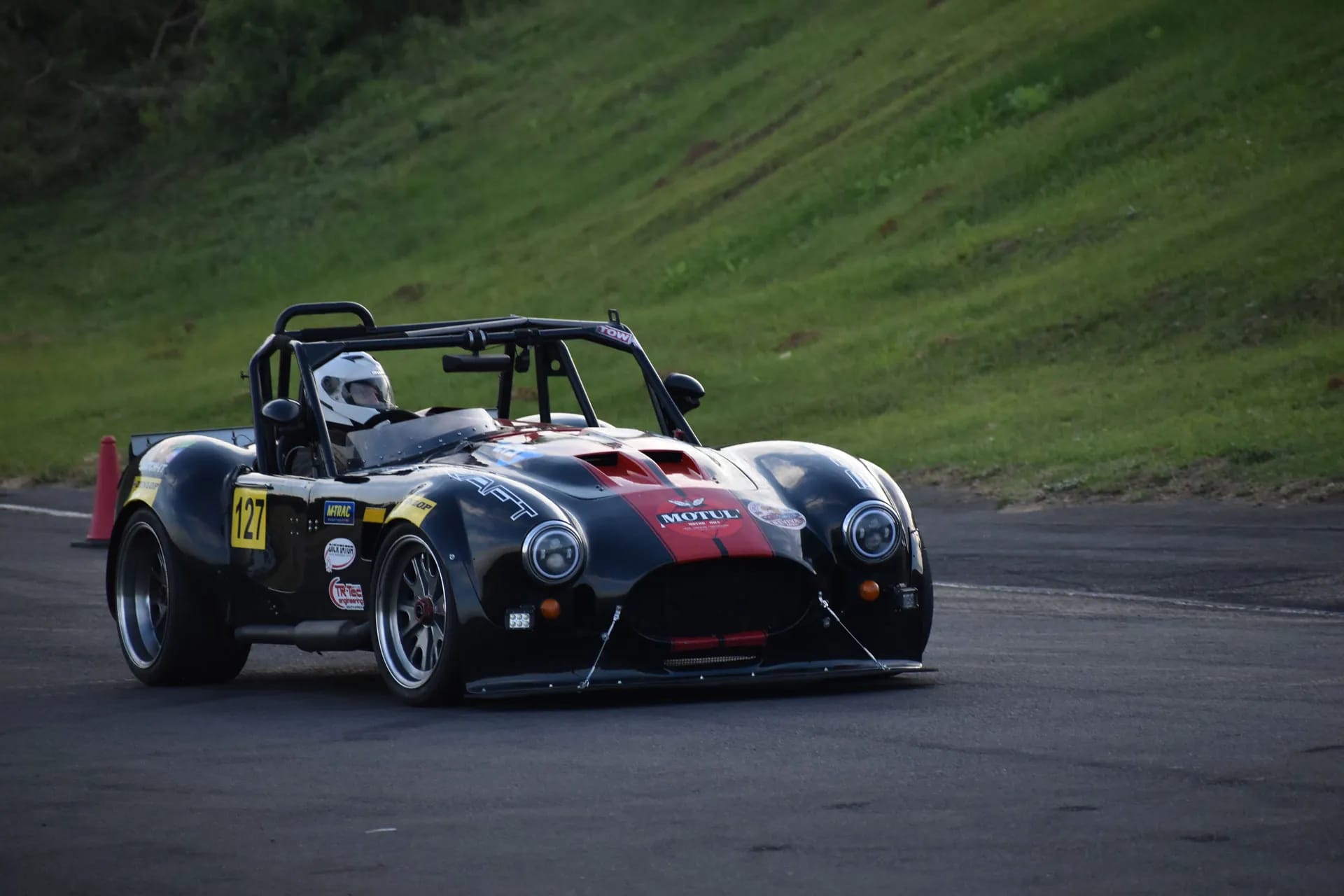 Black and red open-cockpit race car with driver driving on an asphalt track next to a green grassy slope.
