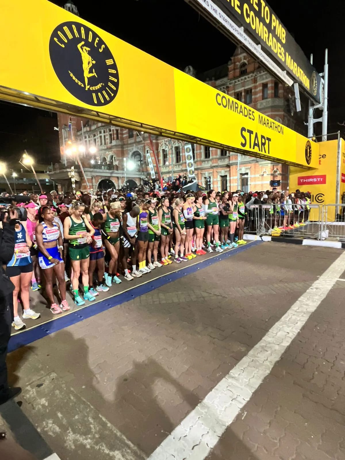Elite runners stand at the Comrades Marathon start line at night, beneath a yellow banner that reads "Comrades Marathon START." A historic building