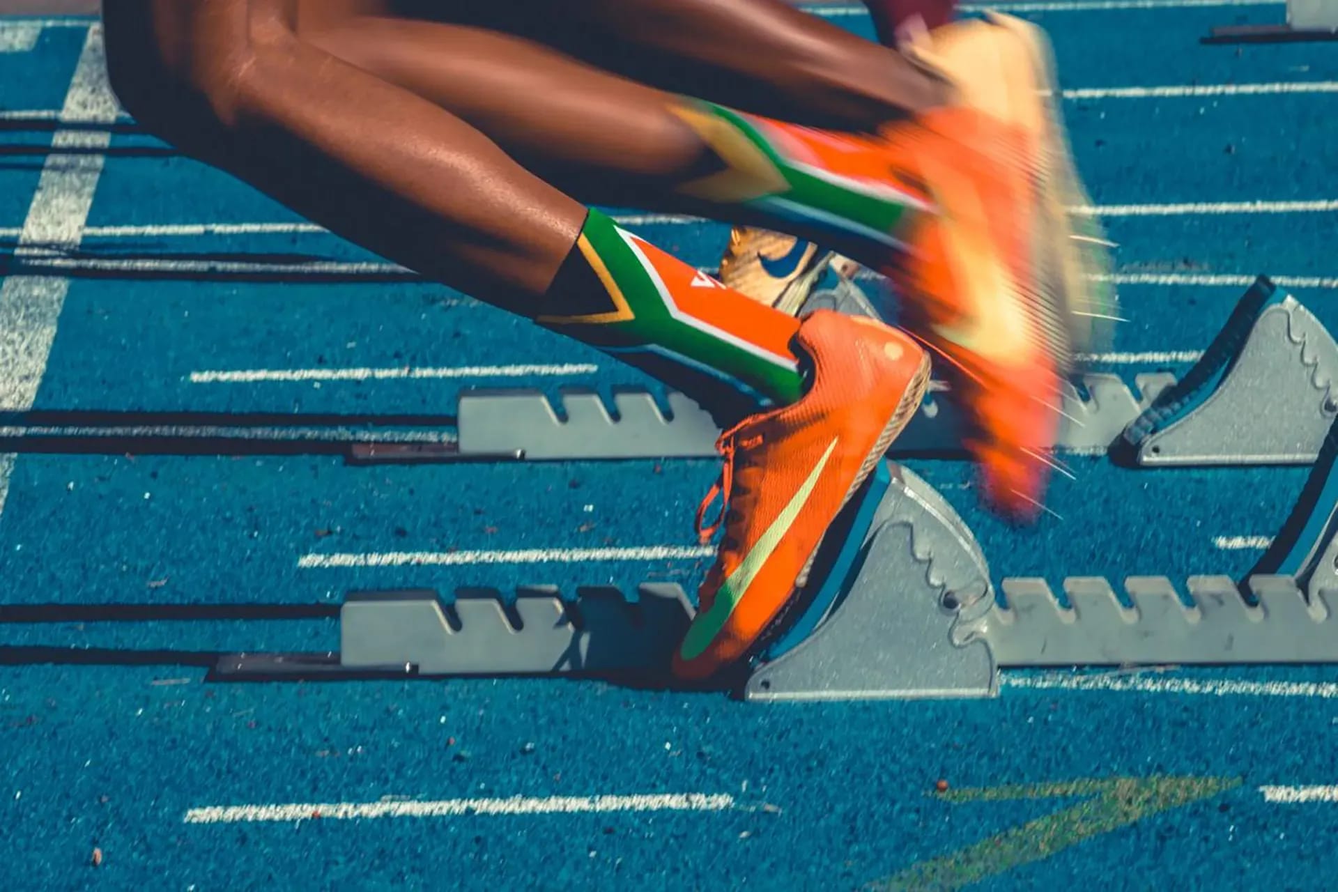 Sprinter in South African flag socks and orange Nike spikes launching from starting blocks on a blue track.