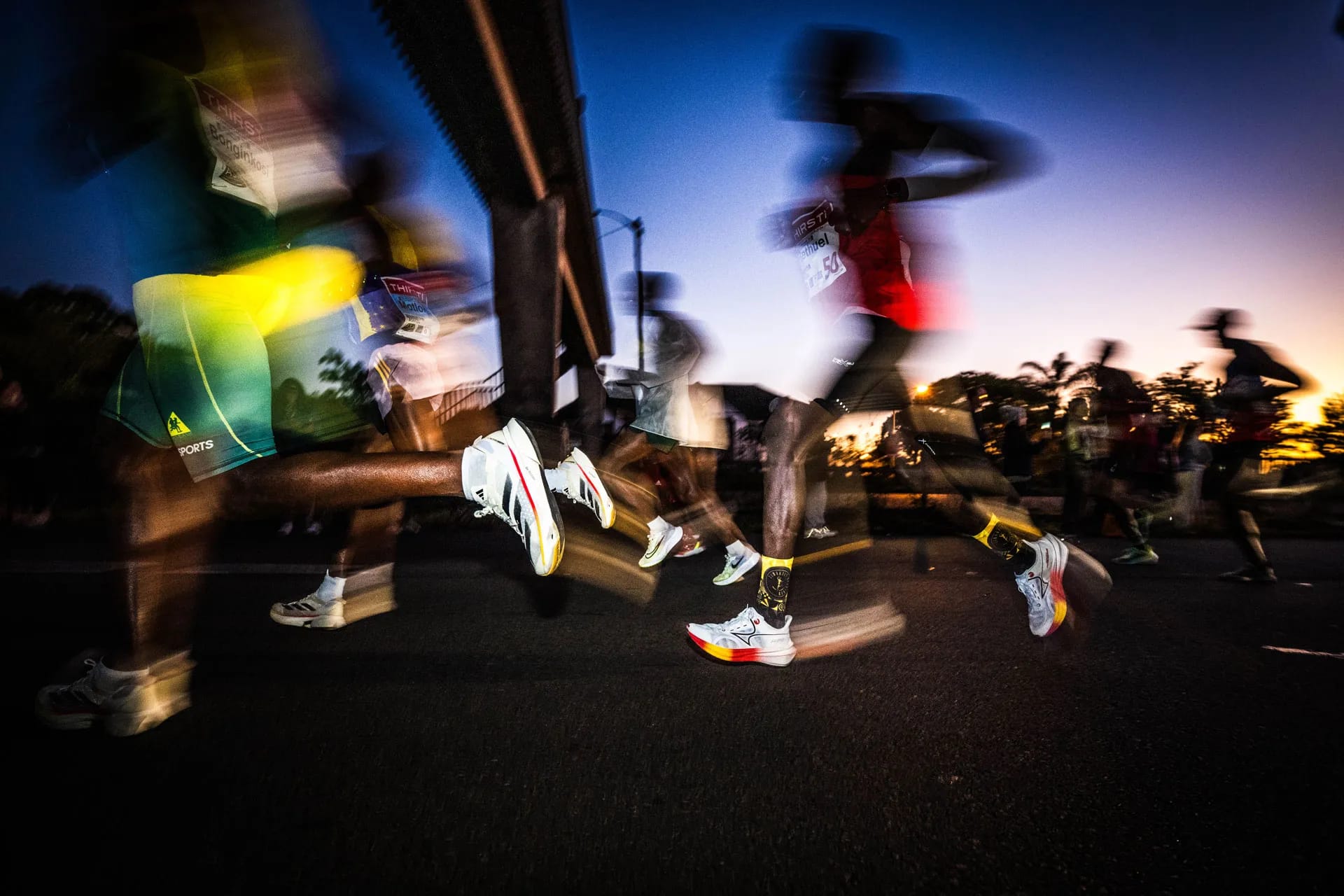 Dynamic low-angle shot of runners in motion blur during a race at dusk. Legs, athletic shoes, and race bibs are visible against a vibrant orange.